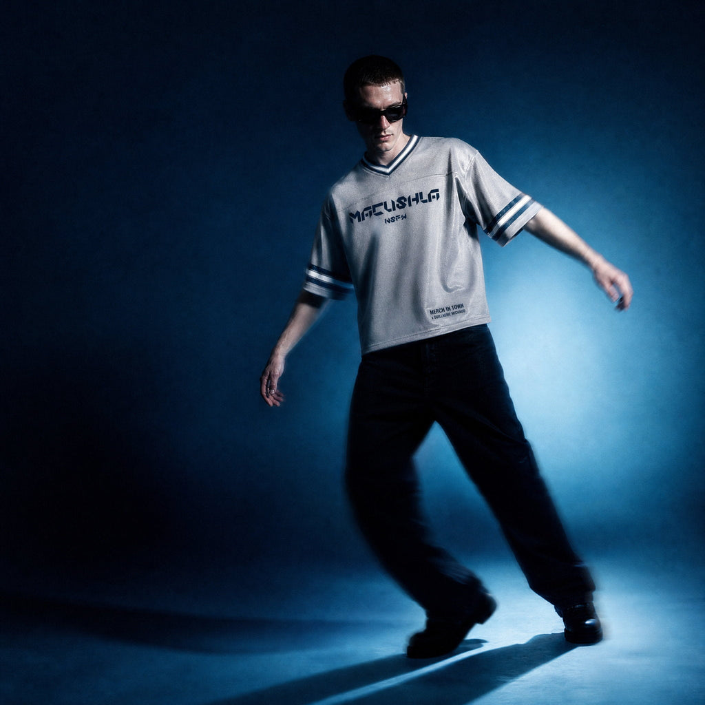 Model in gray boxy striped soccer jersey by Merch in Town x Guillaume Michaud, worn with black jeans and sunglasses under a studio spotlight.