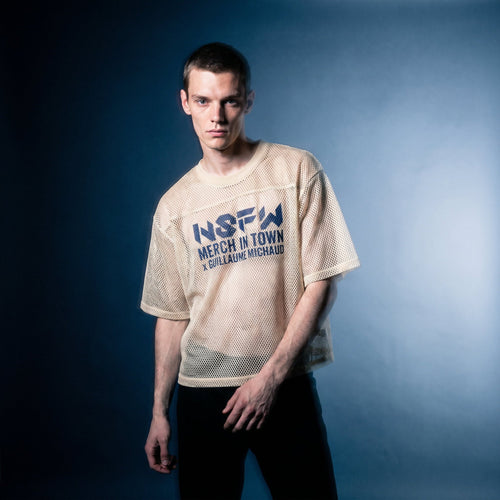 Portrait of the beige mesh NSFW tee by Merch in Town x Guillaume Michaud, worn with black trousers.