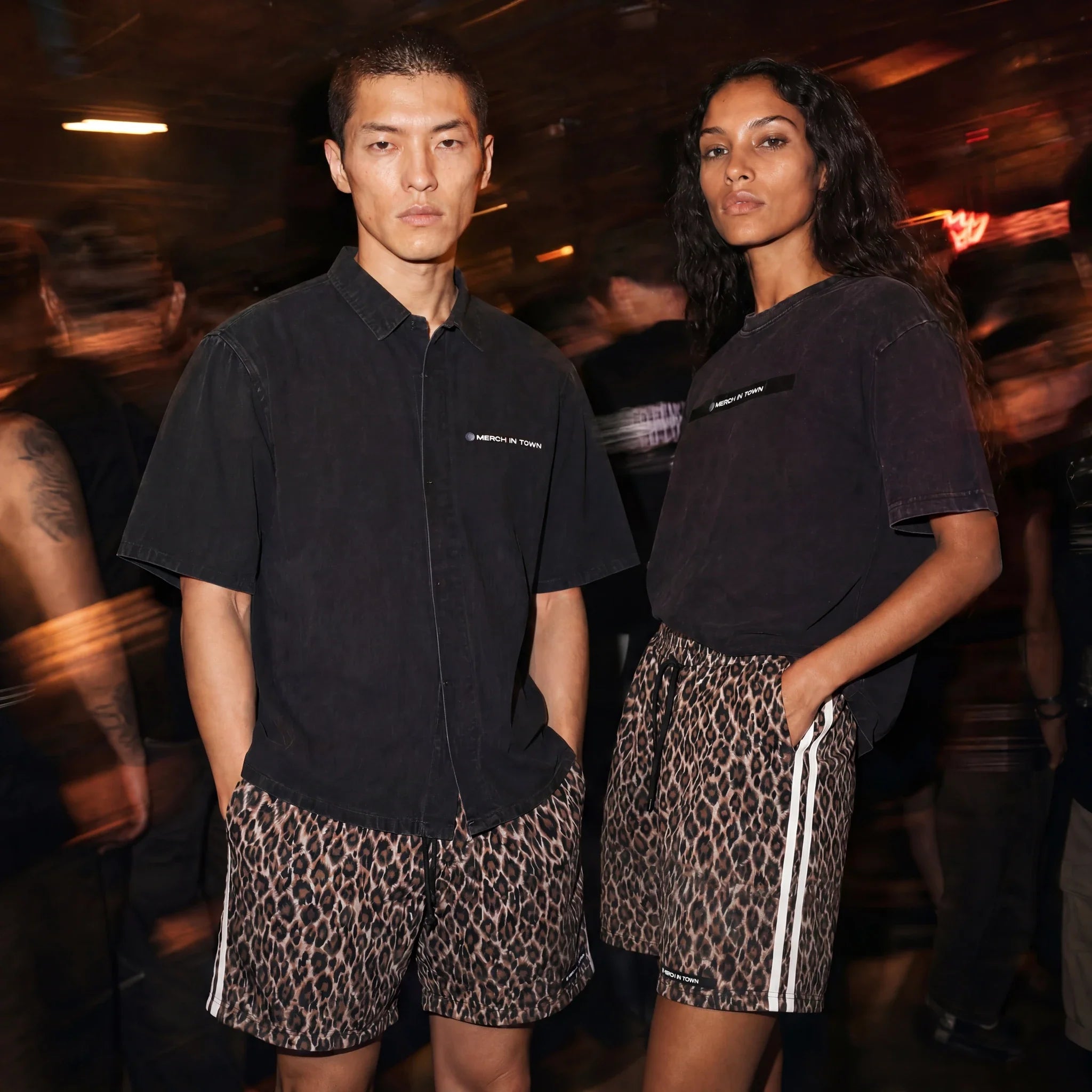 Two models at an underground club, one in the Merch in Town black lyocell shirt, both in leopard drawstring shorts.