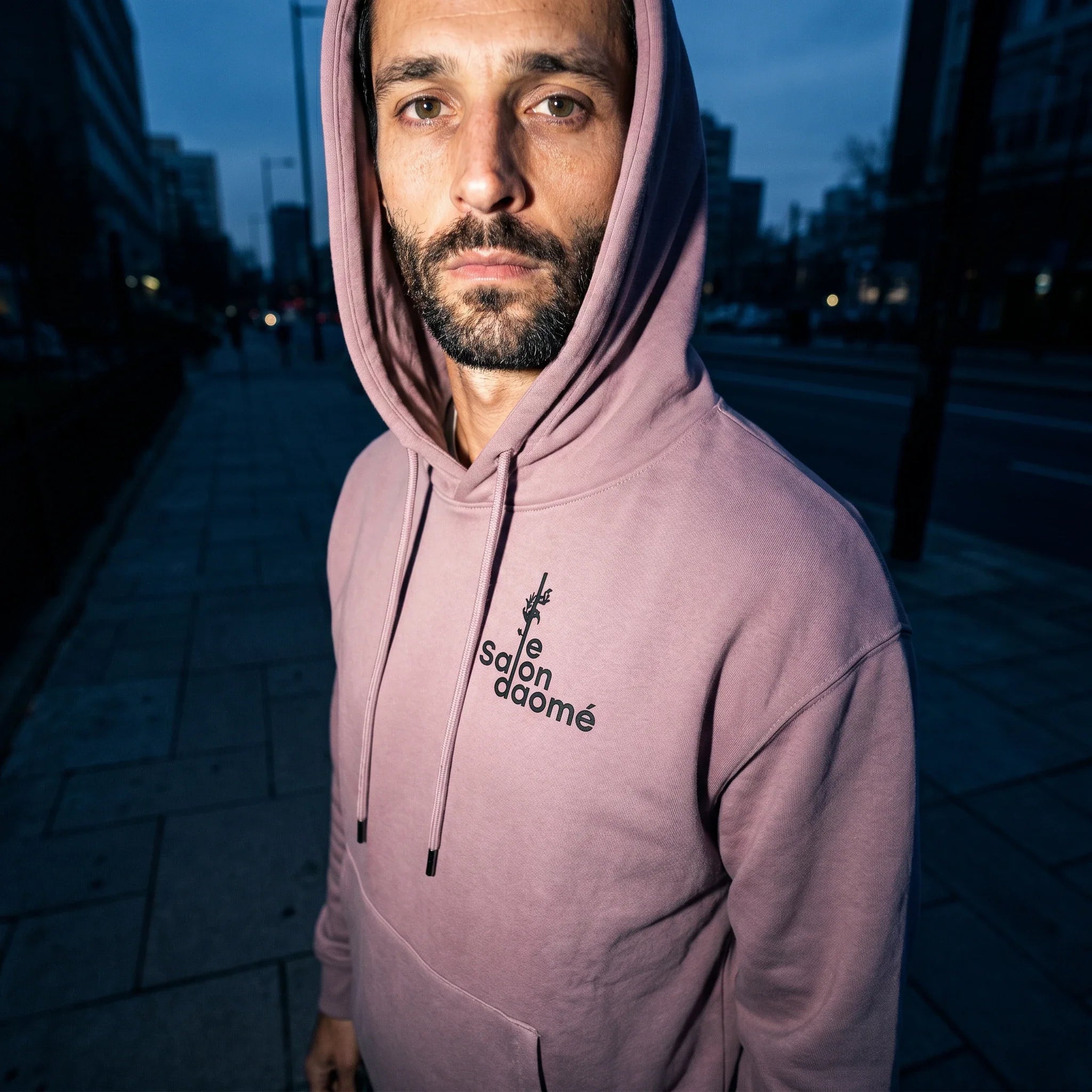 Salon Daomé ash purple fleece hoodie worn with the hood up on an evening street, embroidered chest logo visible.