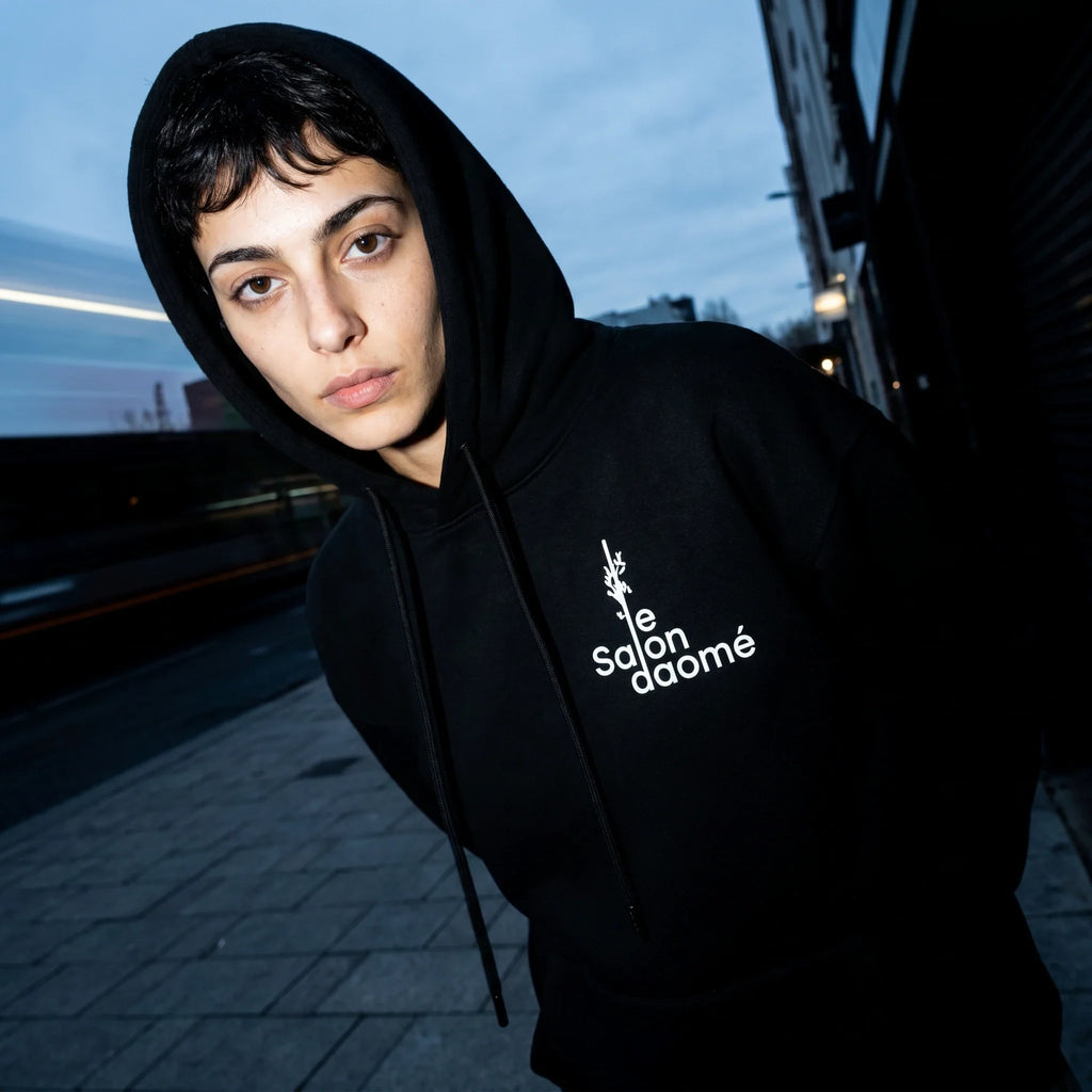 Salon Daomé black fleece hoodie worn with the hood up at dusk, embroidered chest logo visible.
