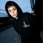 Salon Daomé black fleece hoodie worn with the hood up at dusk, embroidered chest logo visible.
