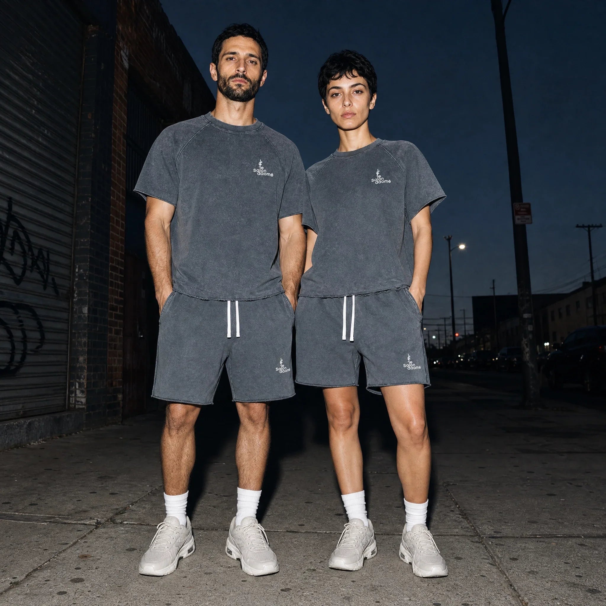 Two people wearing the Salon Daomé mineral wash raw edge t-shirt and shorts set in medium gray on a city street.
