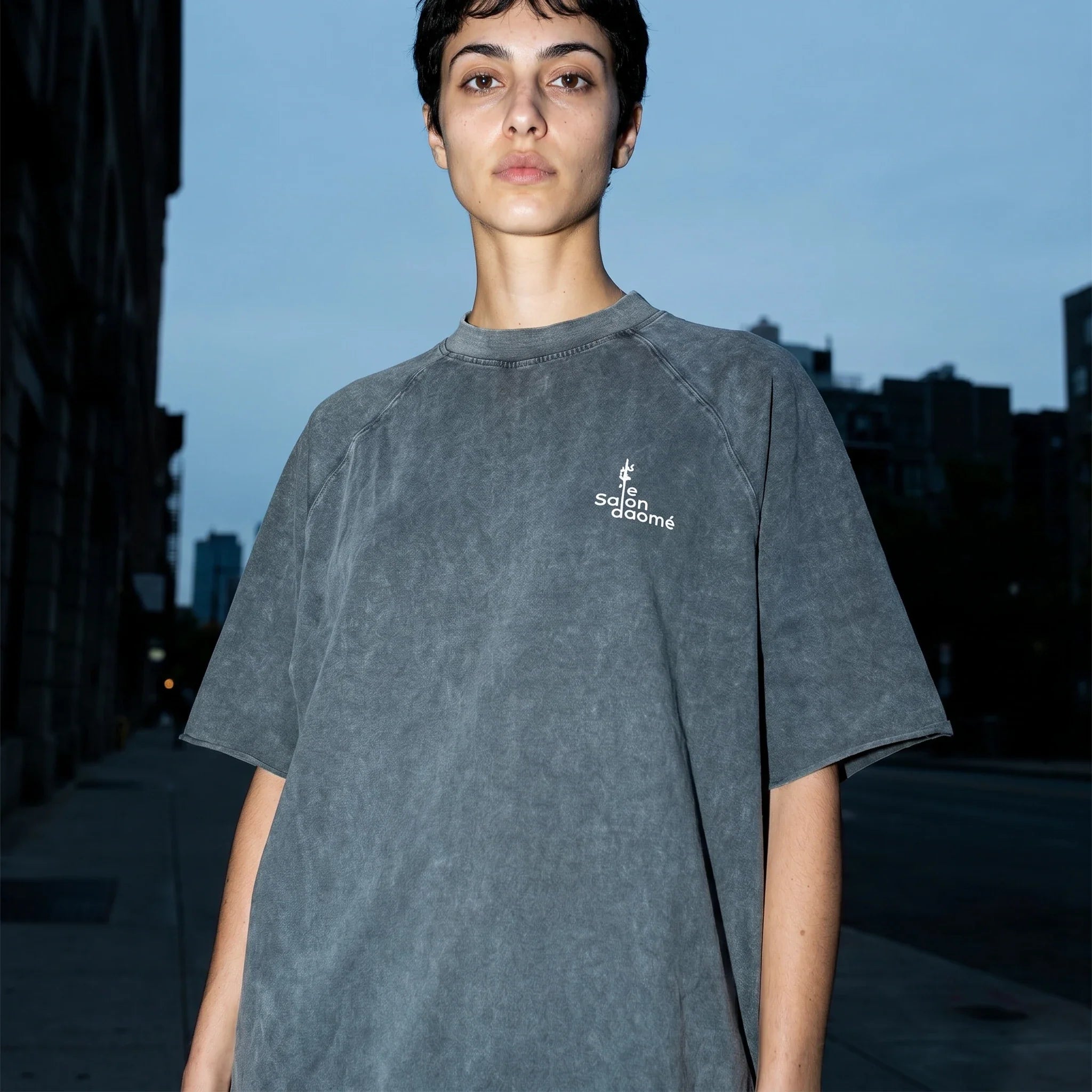Salon Daomé mineral wash raw edge t-shirt in medium gray, oversized fit, on a city street at dusk.