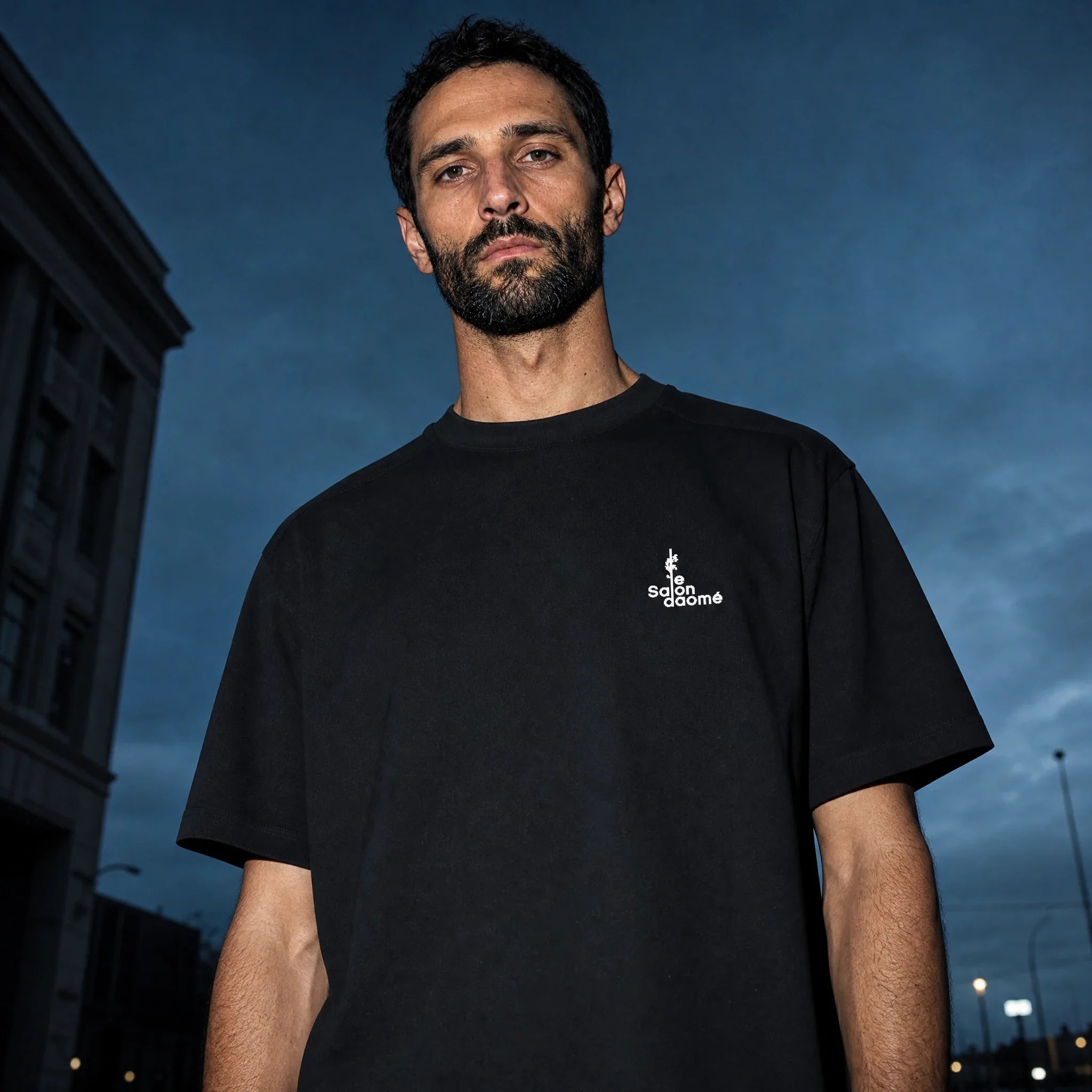 Salon Daomé black Sorona quick-dry boxy t-shirt against a dusk sky, chest logo visible.